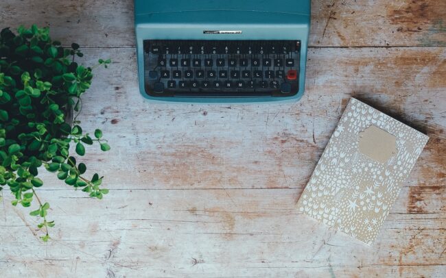 typewriter on a desk next to a notebook and a plant