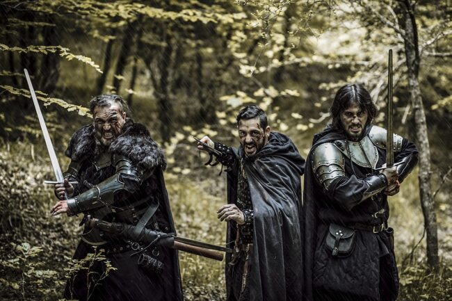 3 men in medieval clothes holding swords, grimacing and looking menacing in the woods